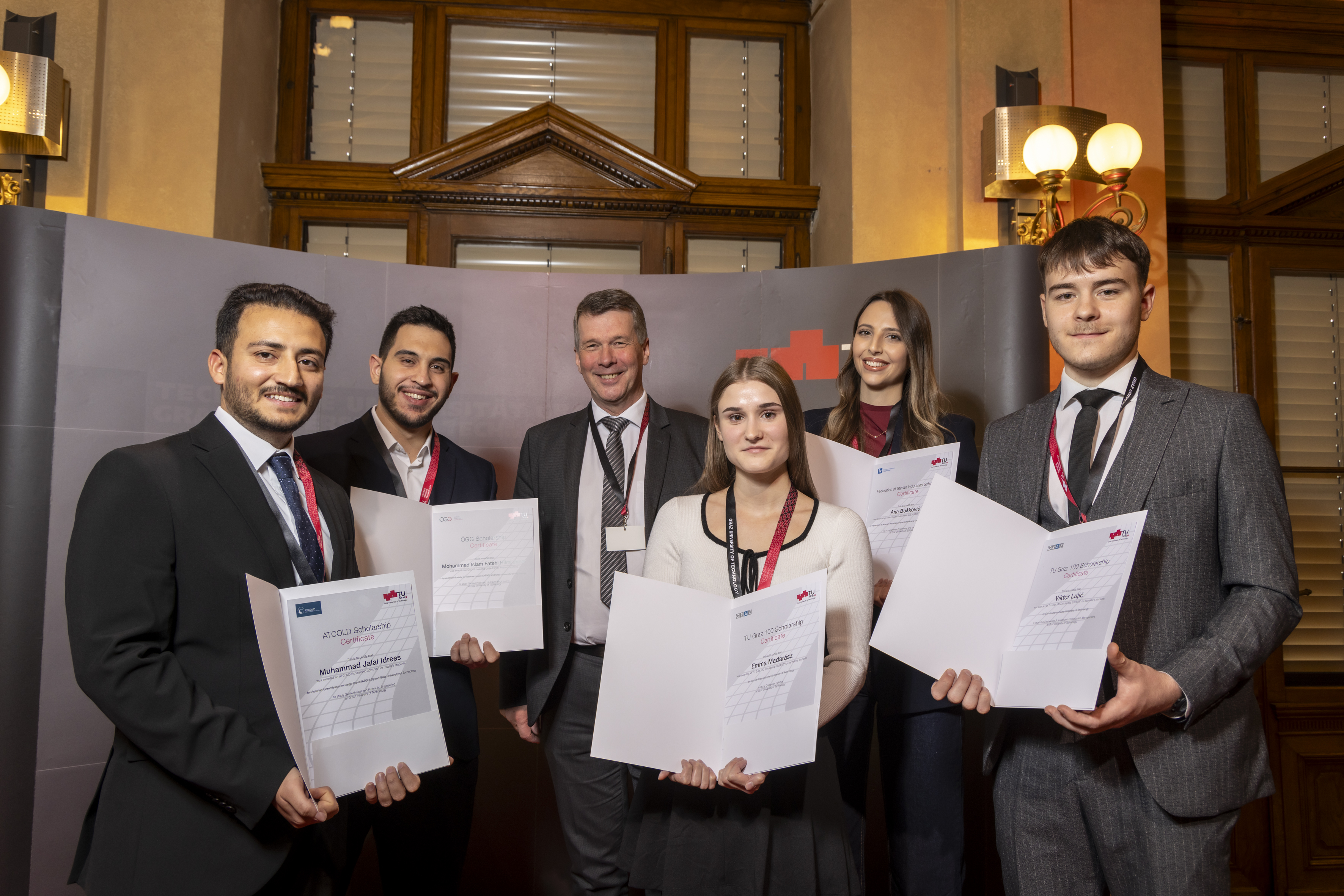 Six people, four men and two women, smile into the camera. Three of the men and the two women are holding certificates recognising them as scholarship recipients.
