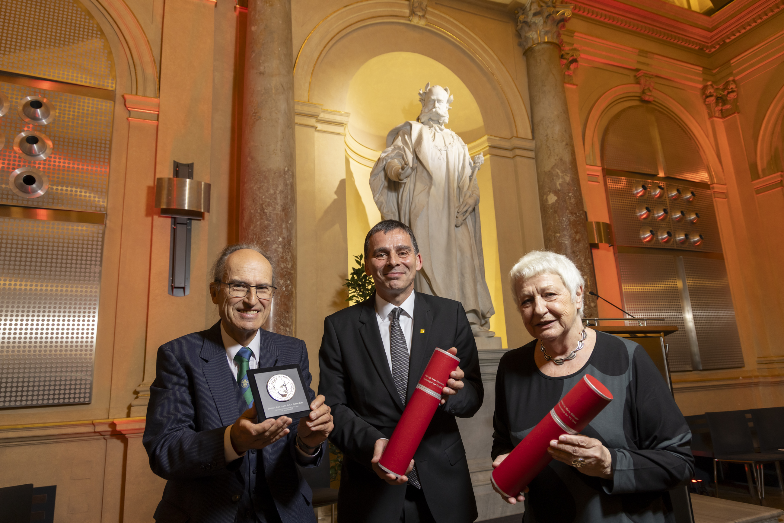 Eine Frau und zwei Männer, zwei mit Urkundenrollen in den Händen, einer mit einer Medaille in einer Schatulle.