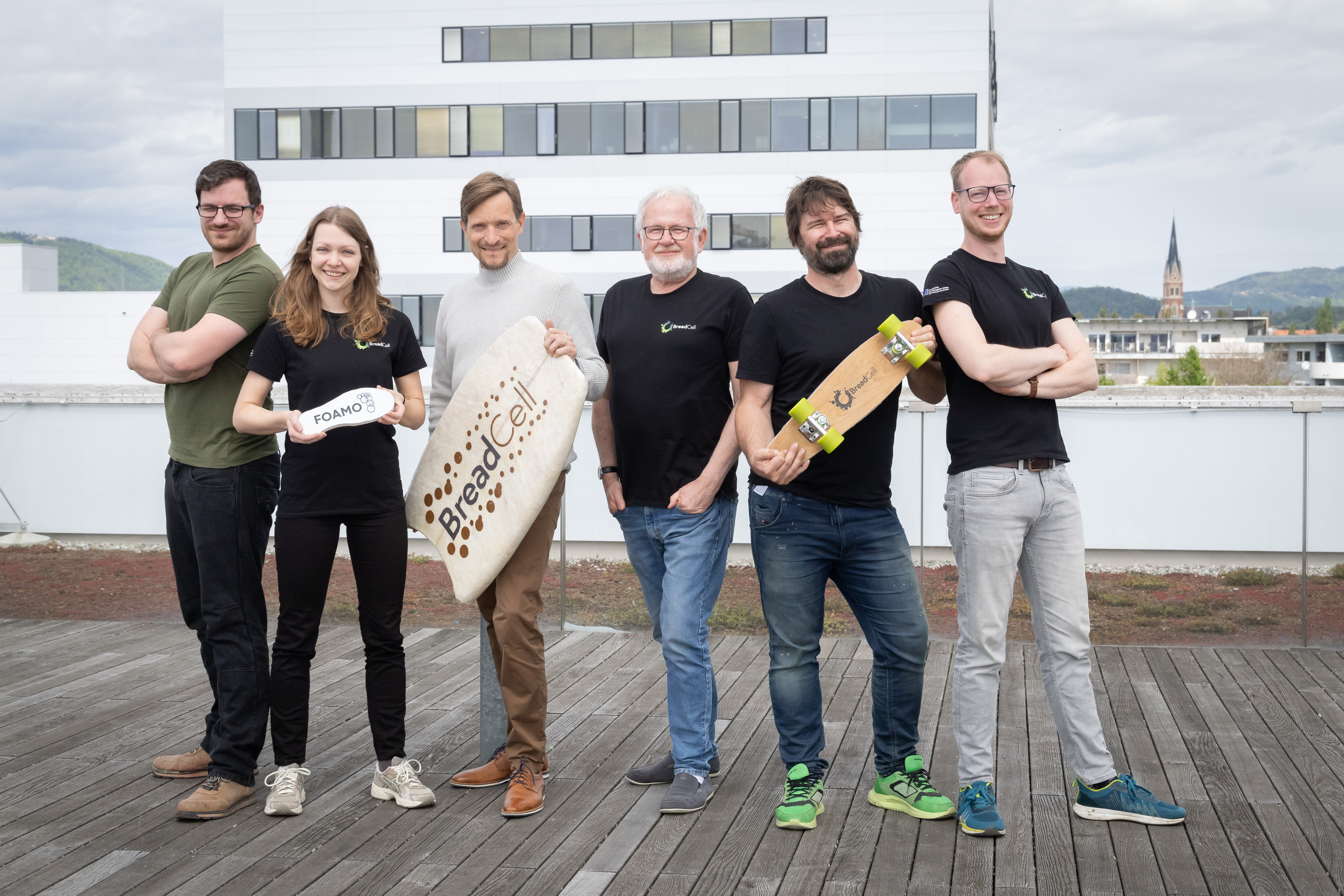Six people are standing next to each other, five men and one woman. The woman is the second person from the left and is holding a shoe sole labelled ‘FOAMO‘. The third person from the left is holding a bodyboard labelled ‘BreadCell’. The second person from the right is holding a skateboard labelled ‘BreadCell’.