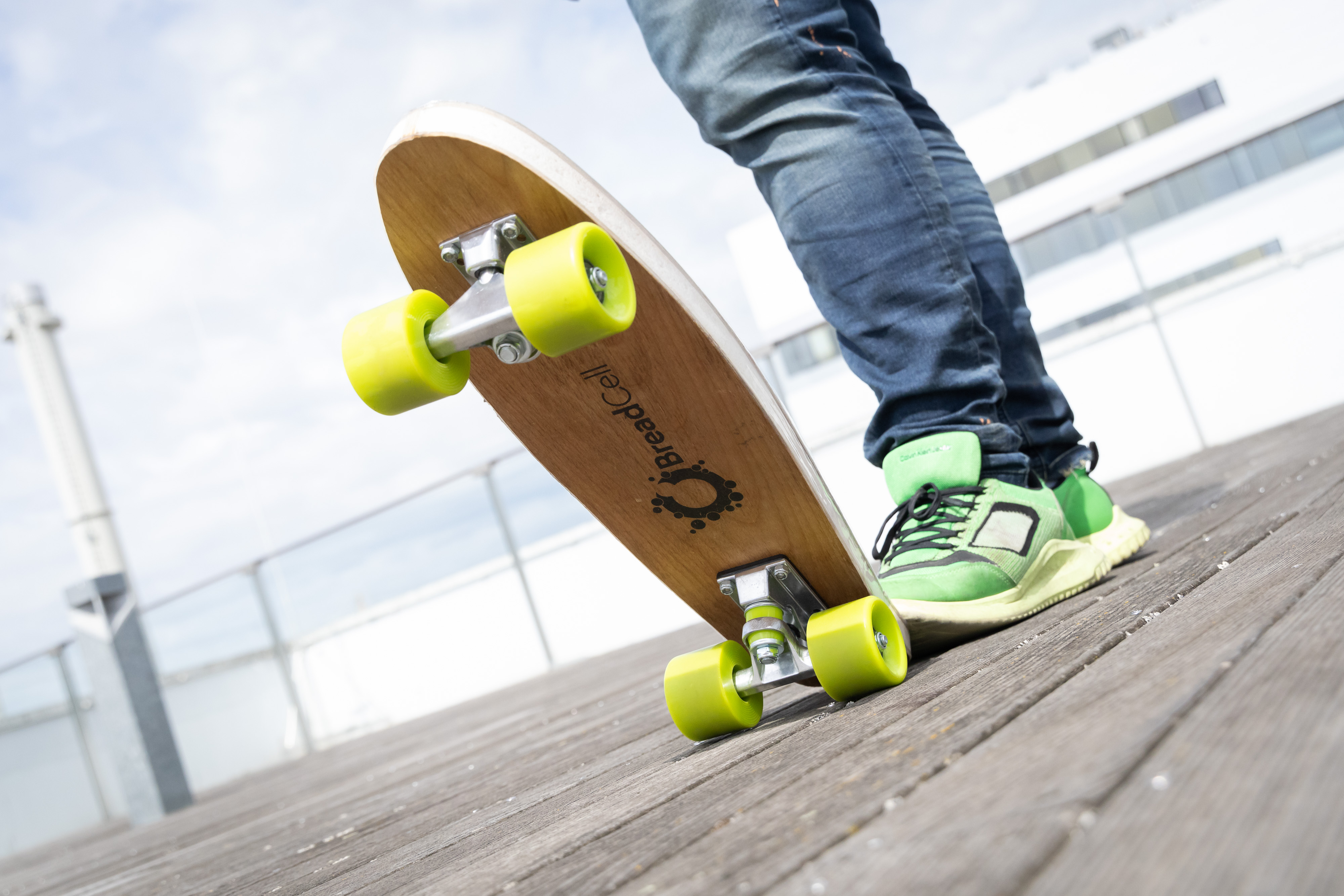 A skateboard with green wheels, the end of which someone is pushing down with one foot so that it is pointing upwards at the front. The word ‘BreadCell’ can be read at the bottom of the skateboard.