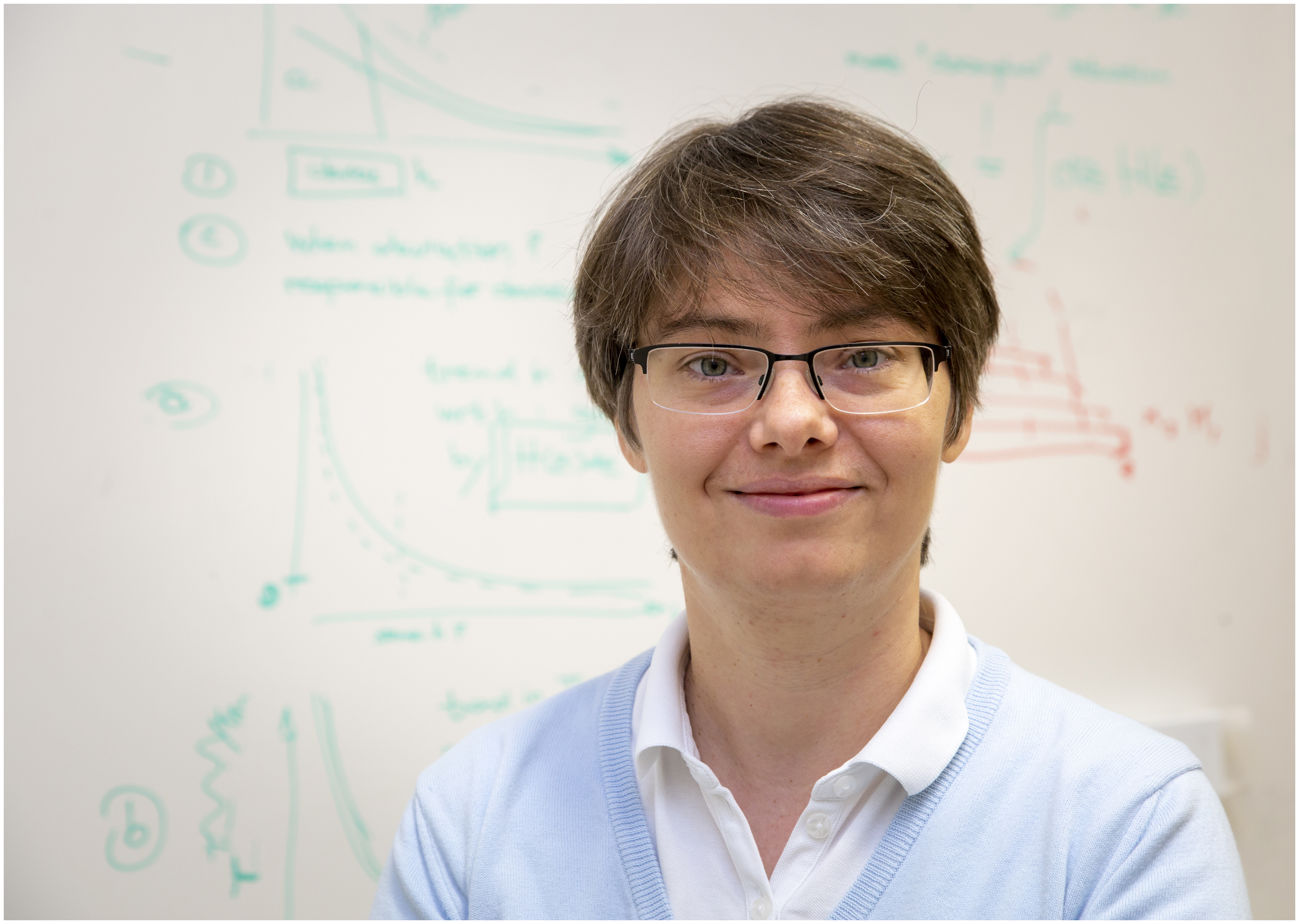 A woman wearing glasses stands in front of a blackboard covered with mathematical formulas and looks into the camera.