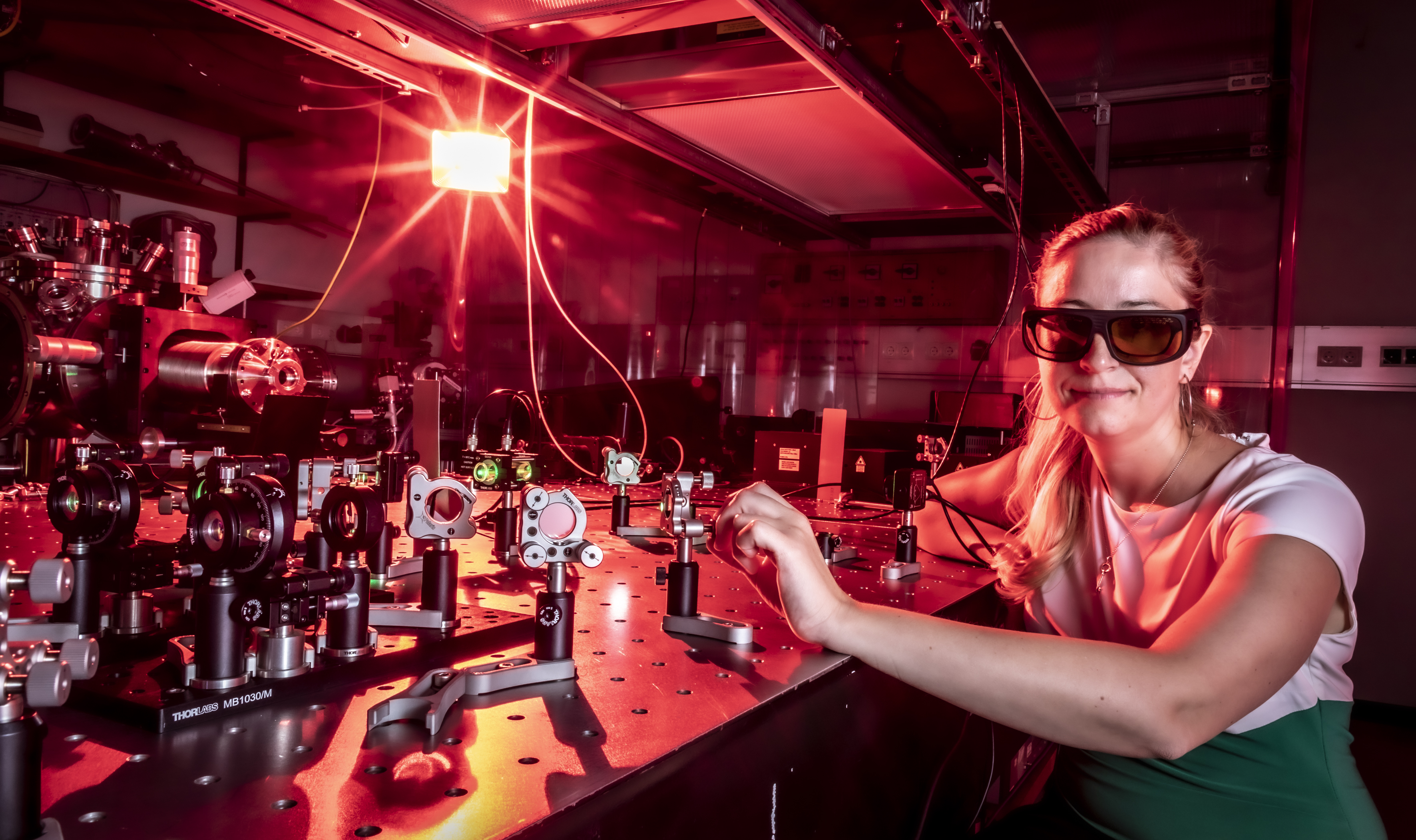 A woman is sitting in a laboratory next to a table with an experimental setup, wearing safety goggles.