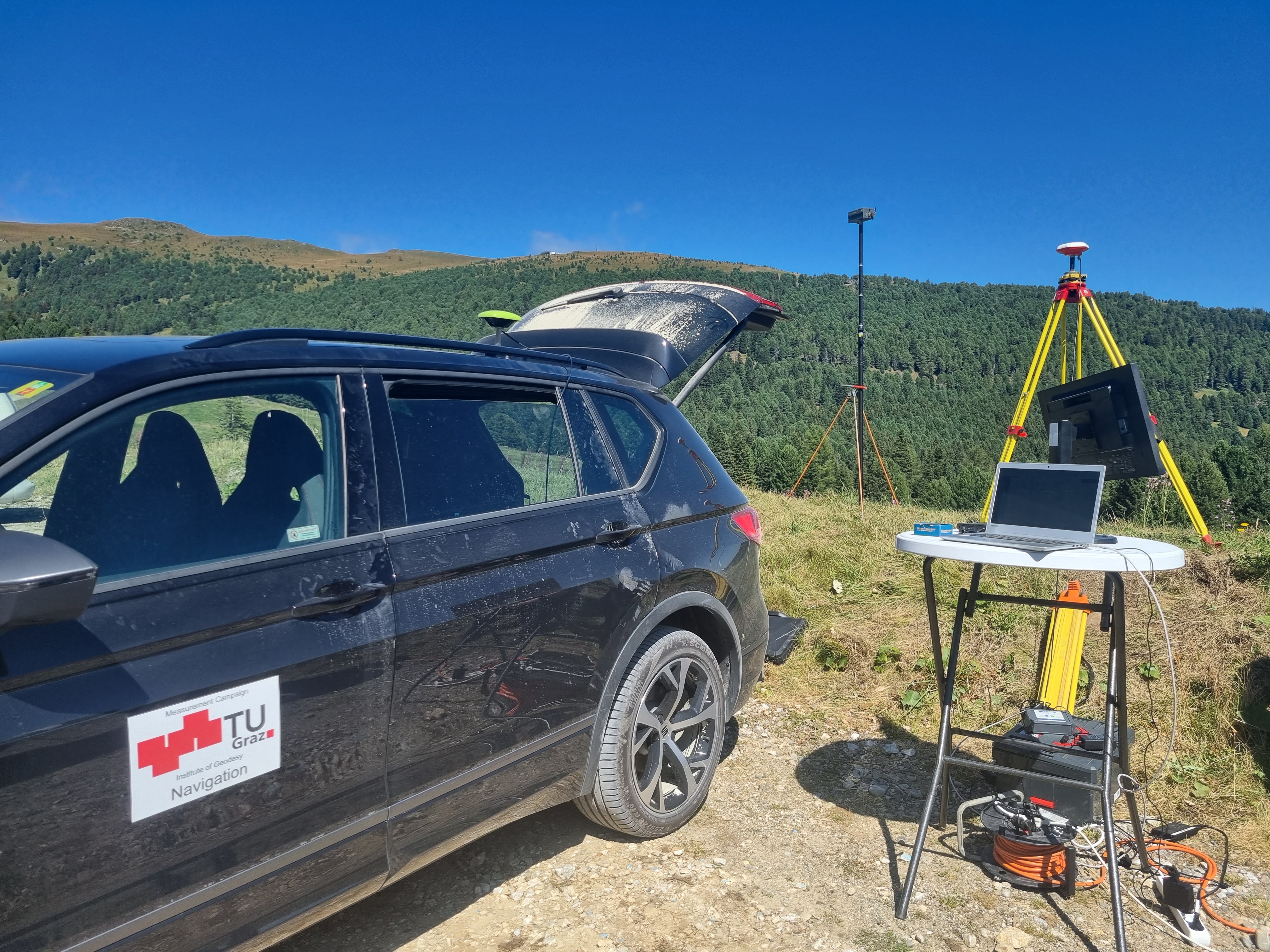 Ein Auto steht neben einem kleinen Tisch, auf dem ein Computer steht. Hinter dem Tisch steht eine GNSS-Antenne.