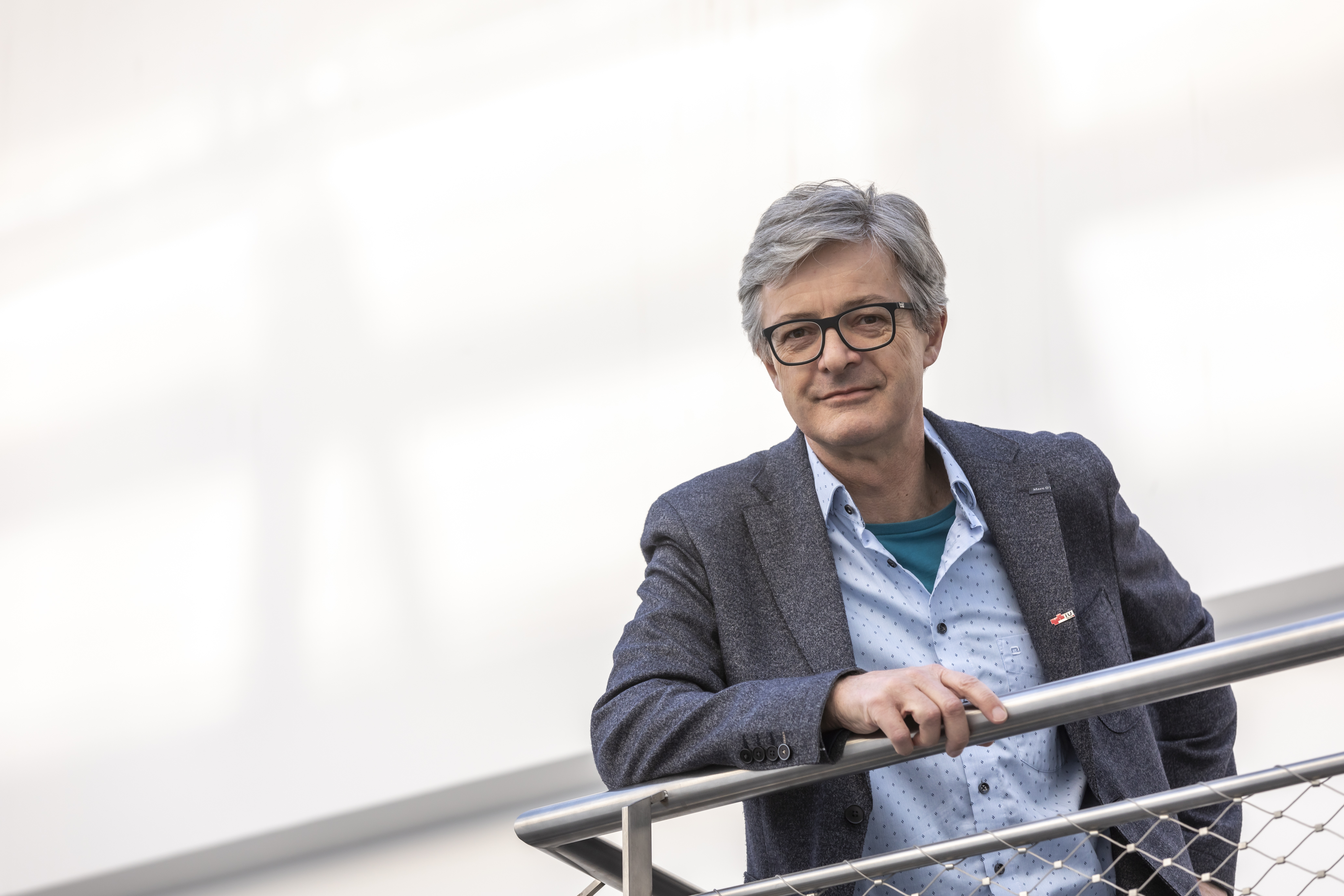 Ein Mann lehnt an einem Geländer und blickt in die Kamera. Es ist nur sein Oberkörper und Kopf zu sehen. Er trägt ein Sakko und ein Hemd.