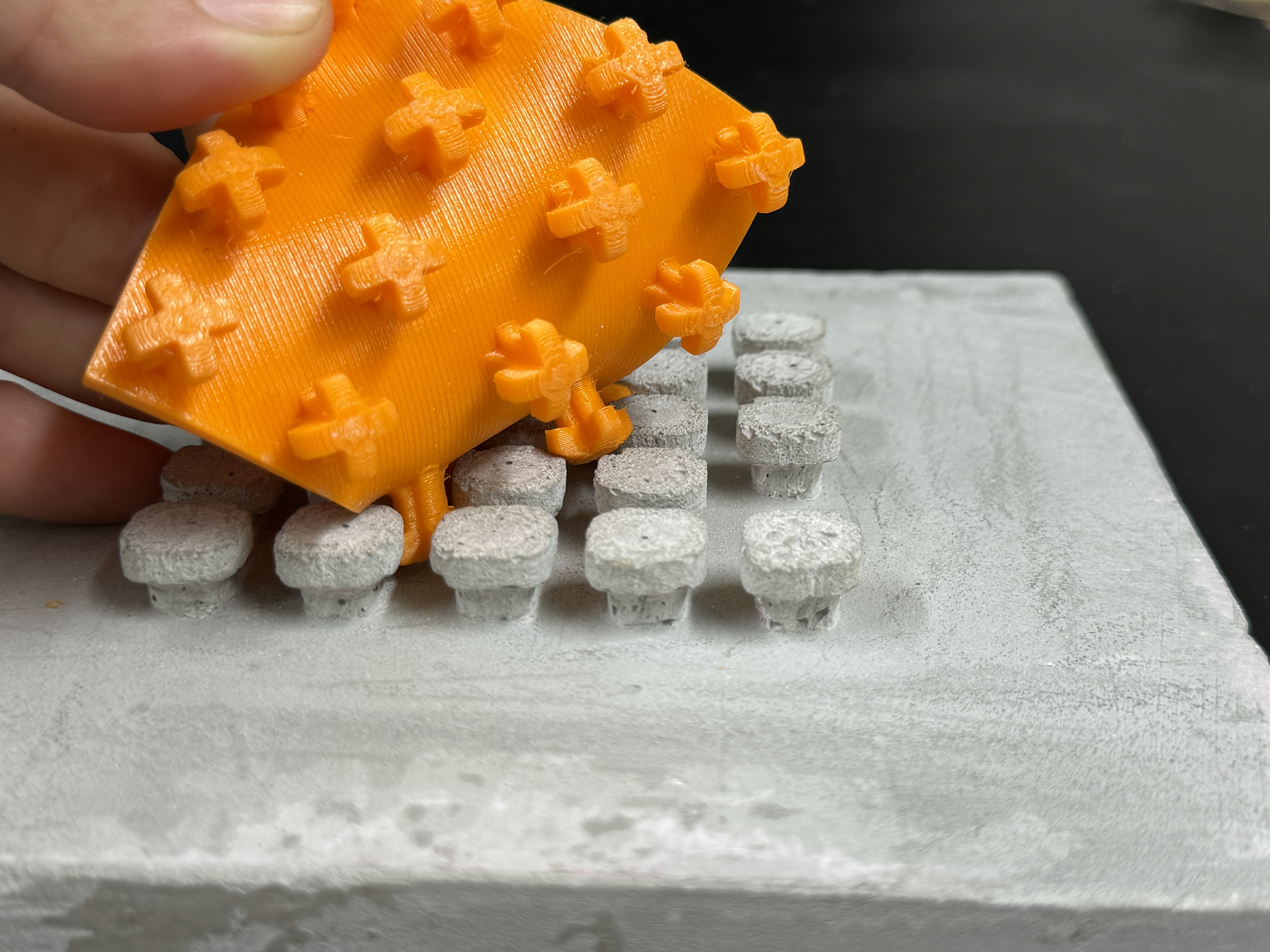 Ein quadratisches, oranges Plastickstück mit Noppen, die sich in ein entsprechendes Gegenstück auf einem Betonteil verhaken.
