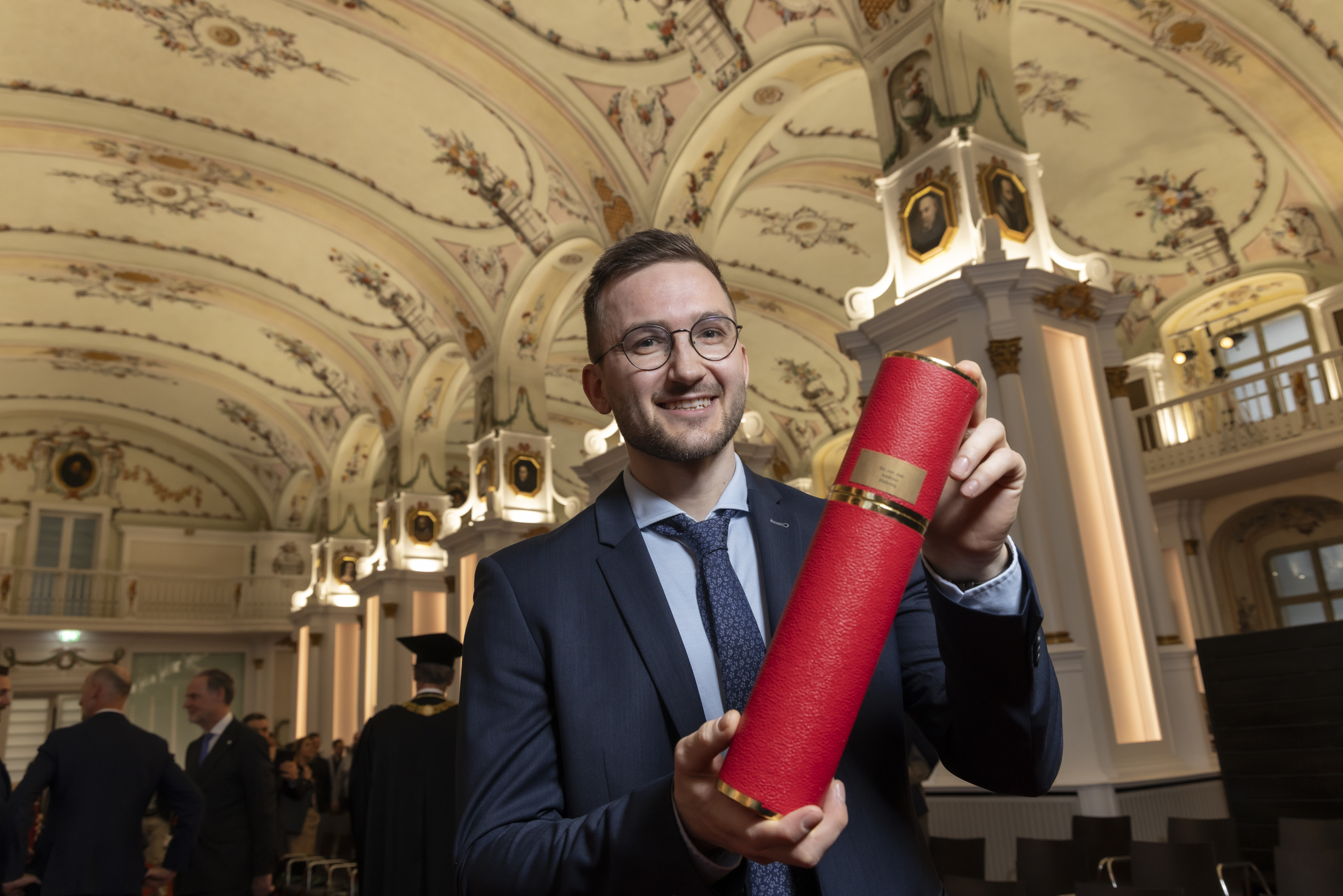 Ein Mann lächelt in die Kamera und hält eine rote Röhre mit seinem Doktoratszeugnis in die Höhe