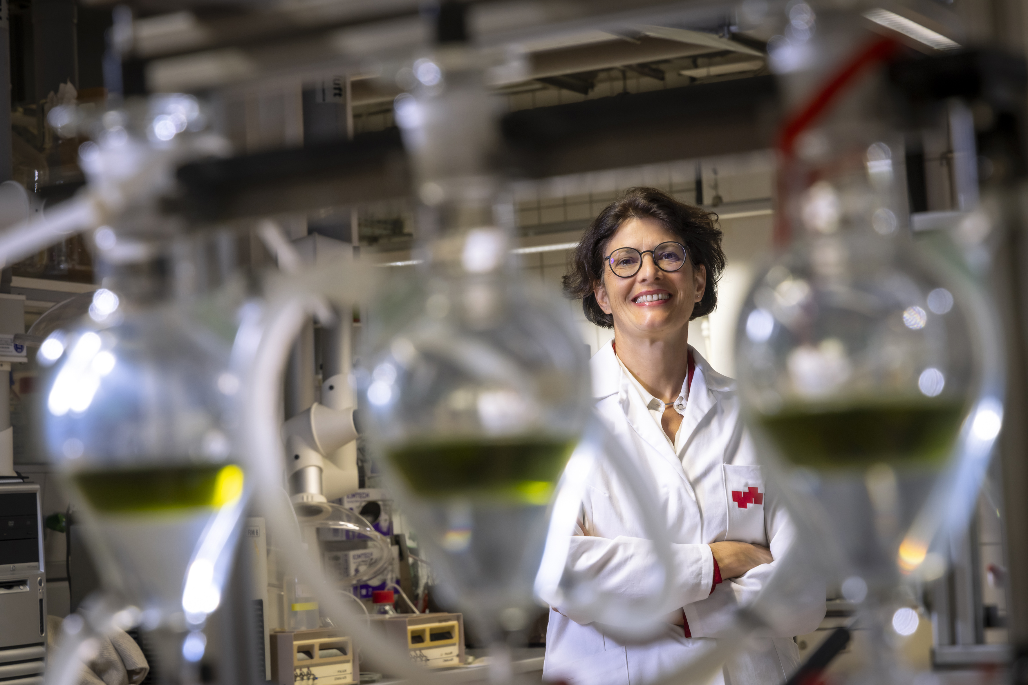 Smiling woman in a lab coat behind glass containers filled with coloured liquid.