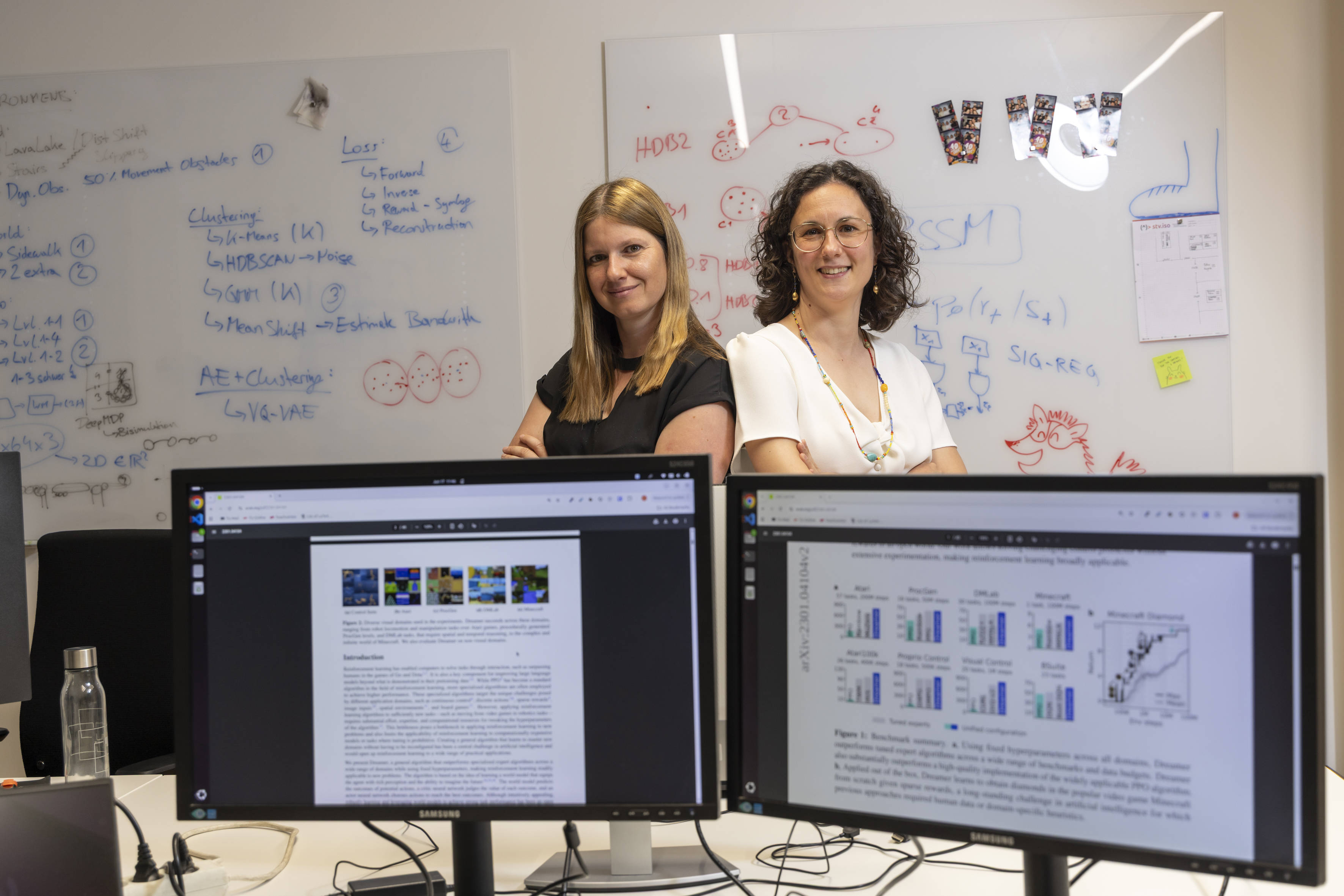 Two women are standing behind computer screens, smiling at the camera.