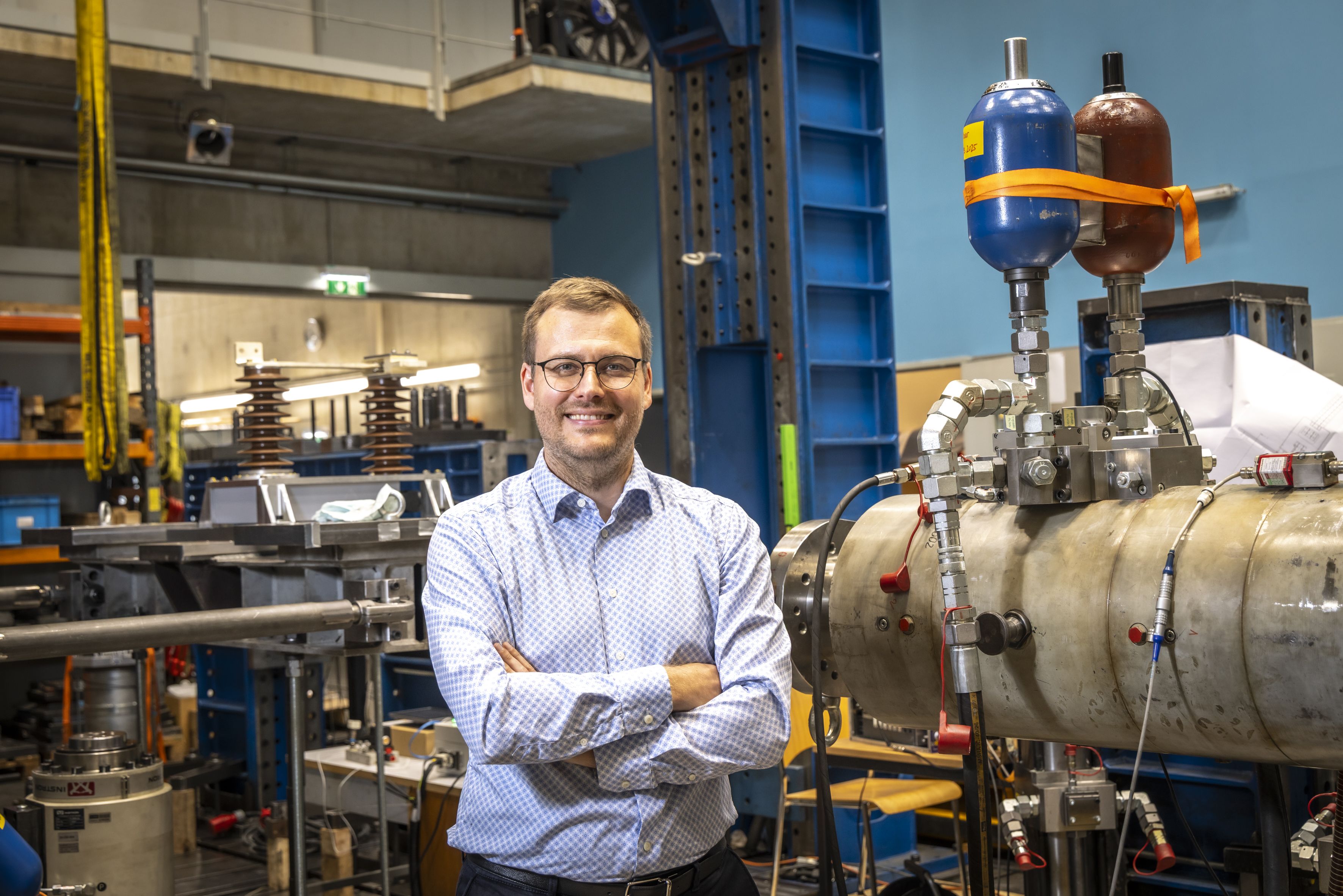 Ein lächelnder Mann mit vor der Brust verschränkten Armen, im Hintergrund eine Halle mit Metallobjekten und Rohren.
