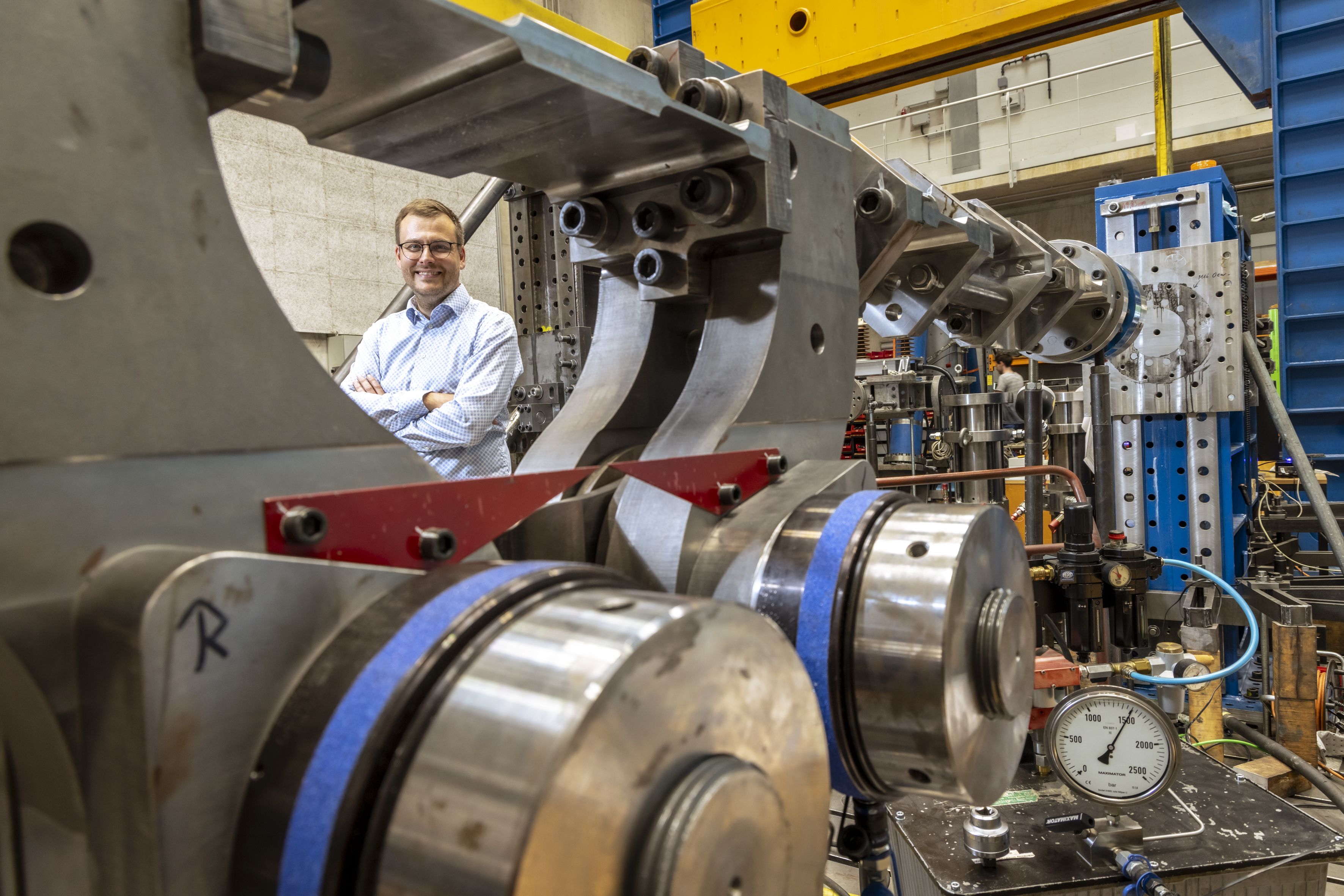 Ein lächelnder Mann mit vor der Brust verschränkten Armen, im Vordergrund ein großes Gebilde aus Metall und Kabeln.