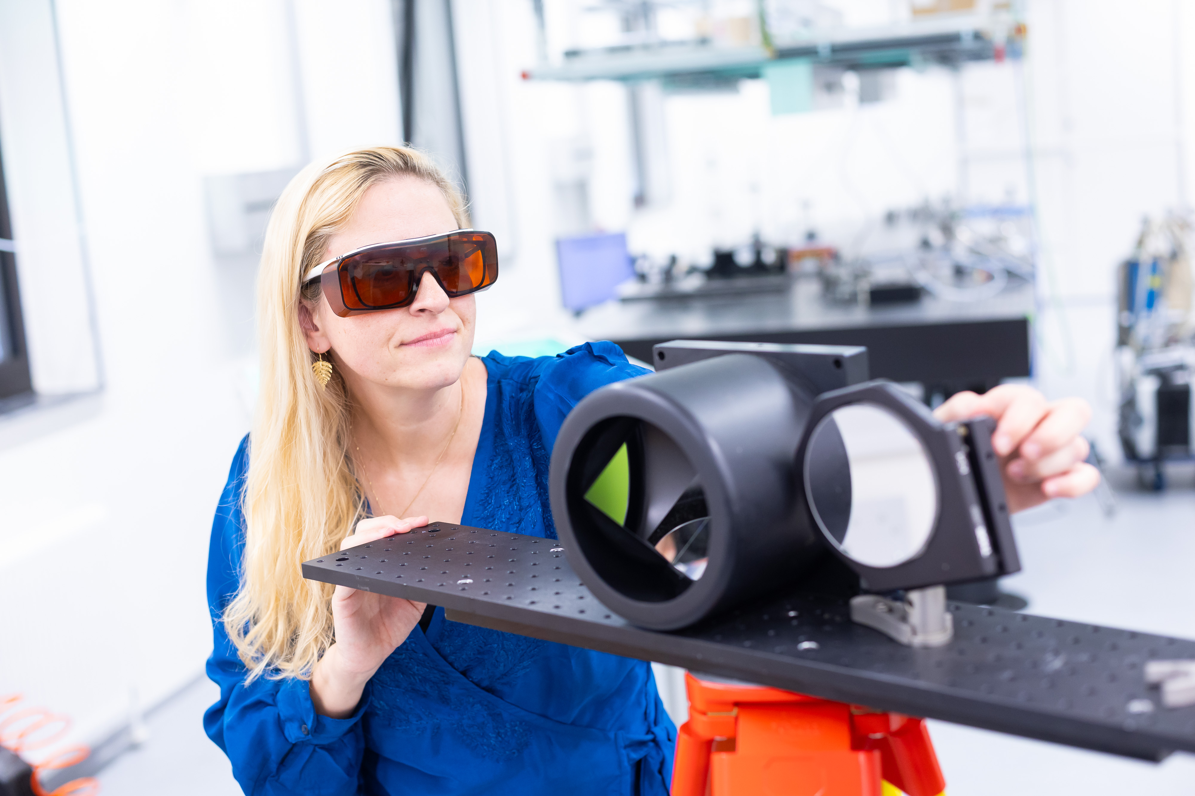 A woman wearing dark safety goggles is looking at a piece of technical equipment in a laboratory.