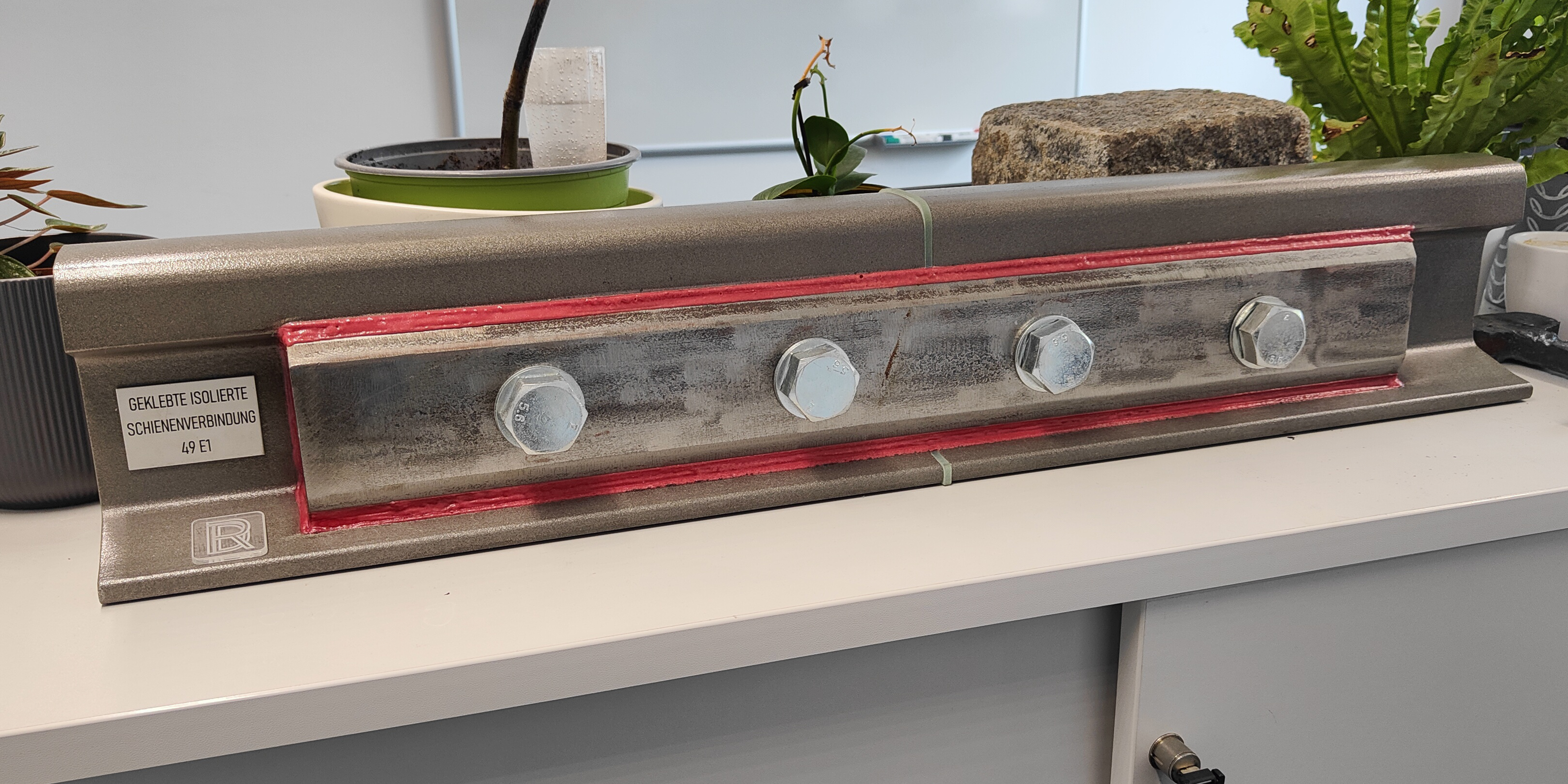 Ein Stück einer Eisenbahnschiene, an deren Seite eine Verstärkung mit Schrauben angebracht ist. In der Mitte ist die Schiene durch ein dünnes Kunststoffteile in zwei Hälften geteilt.