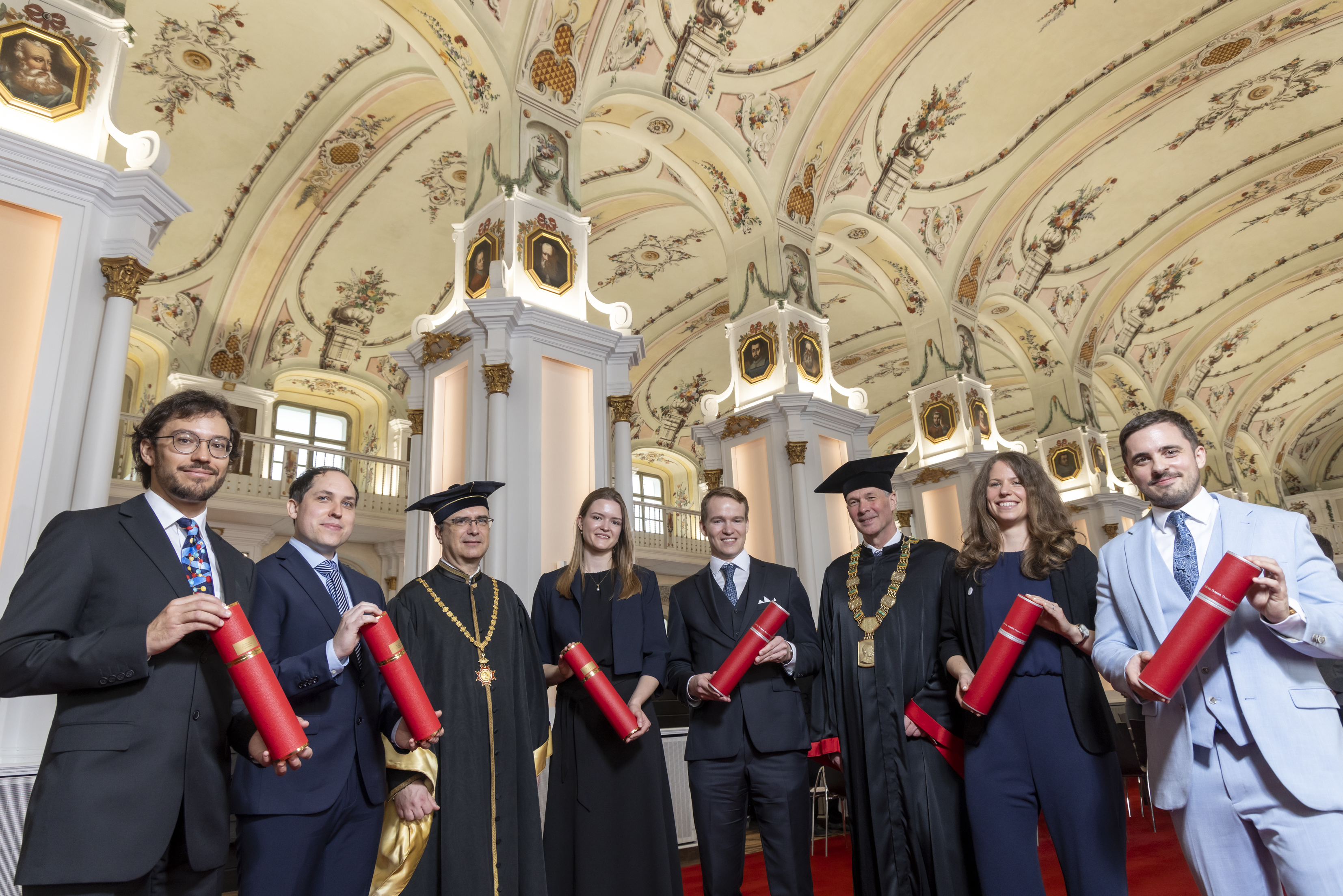 Acht Personen in festlicher Kleidung blicken in die Kamera und lächeln. Sechs von ihnen halten rote Rollen in der Hand.