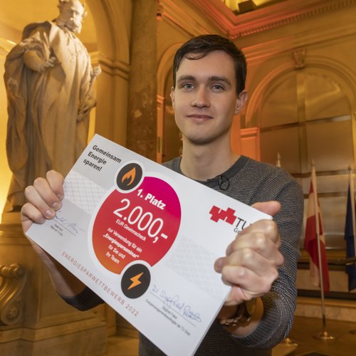 A young man with short, dark hair holds a white cardboard box up to the camera. On it is a red circle with the inscription "1st place".