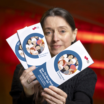 A woman holds three flyers up to the camera. One reads: TU Graz is breaking down barriers!