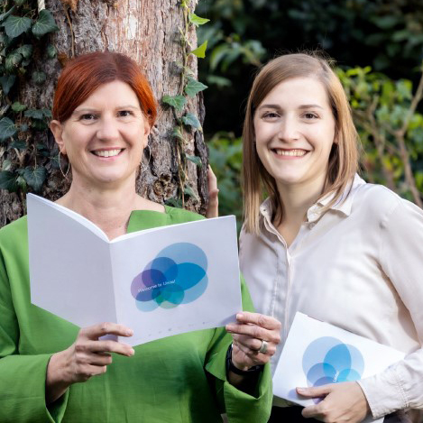 Eine Frau in einer grünen Bluse und eine Frau in einer weißen Bluse stehen vor einem Baum. Sie halten weiße Broschüren in den Händen.