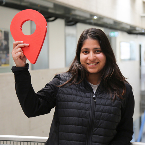 A young woman with long, dark hair and wearing a black jacket holds a red location pin in her right hand.