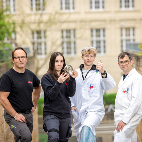 Three men and a woman are standing next to each other. Two of the men are wearing white lab coats, while the woman and the other man are wearing black T-shirts. The coats and T-shirts are printed with the TU Graz logo.