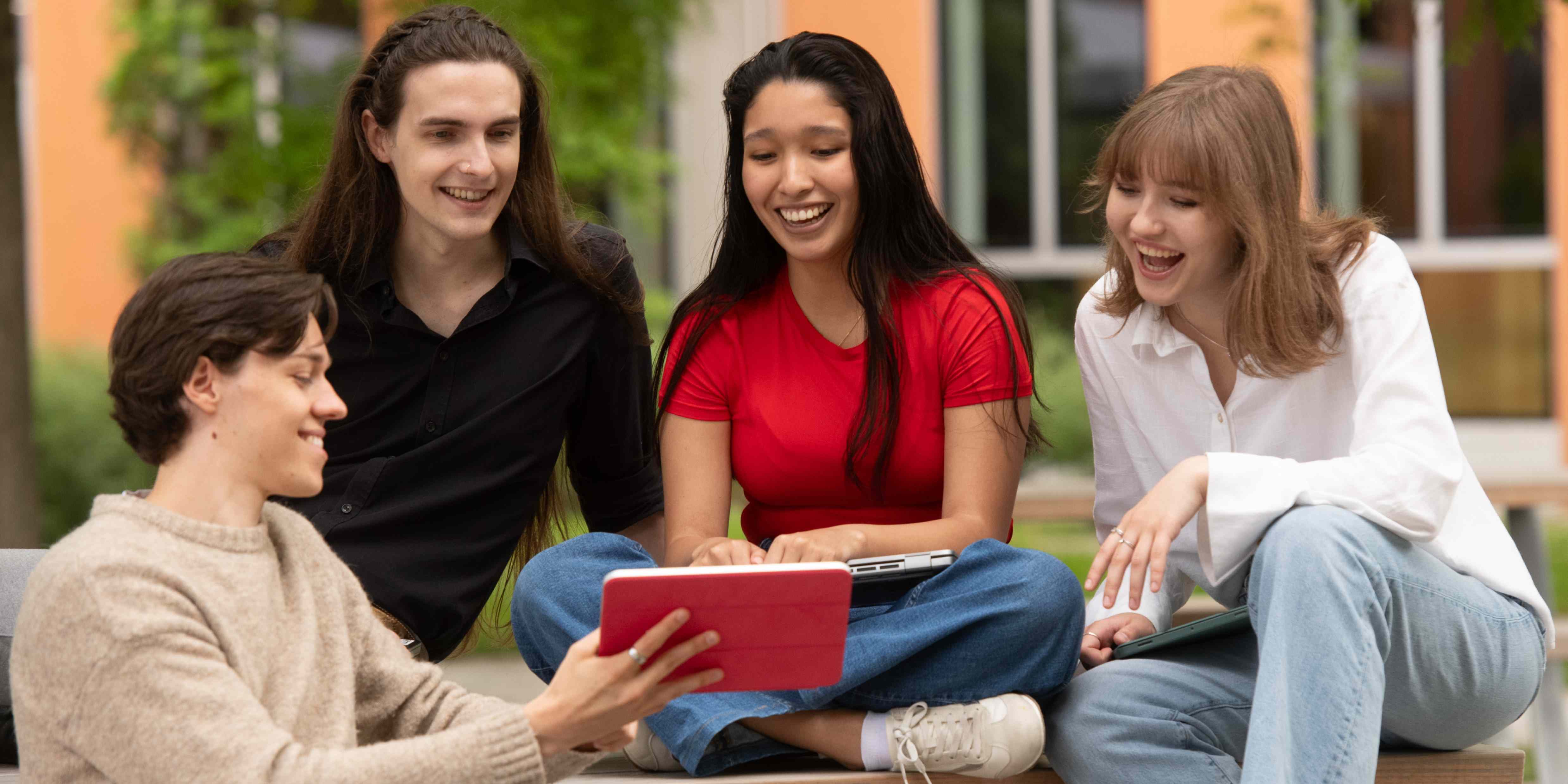 Four students are sitting at the campus, look at computers while chatting.