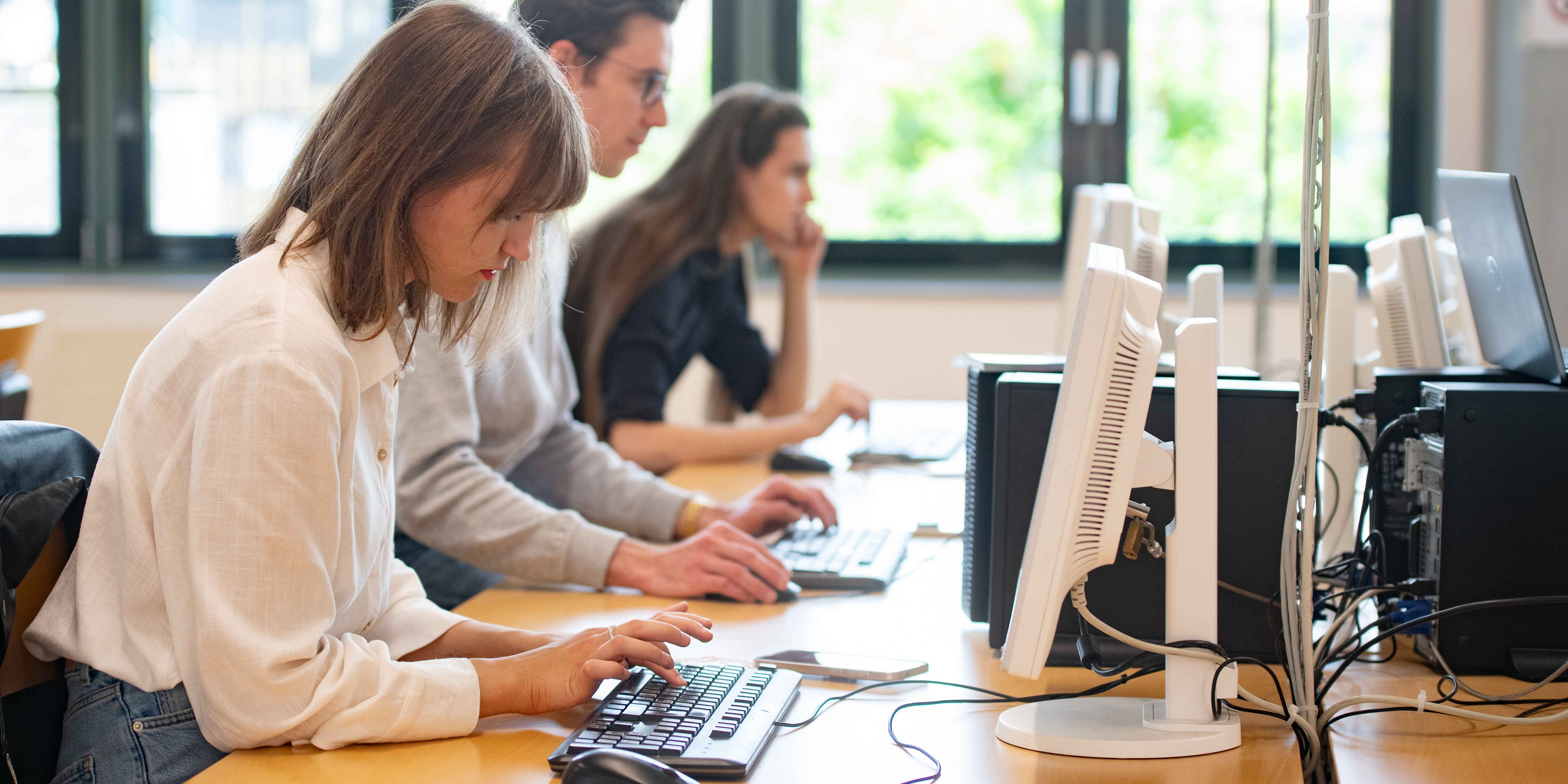 Three students are working on computers at a long table.