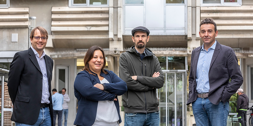 Drei Männer und eine Frau stehen vor einem Gebäude und blicken in die Kamera