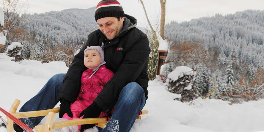 Jürgen Zechner mit gestreifter Strickkappe und schwarzer Winterjacke und seine entzückende kleine Tochter im pinken Skioverall und mit rosa Haube auf einer Rodel im Schnee. Im Hintergrund verschneiter Wald.