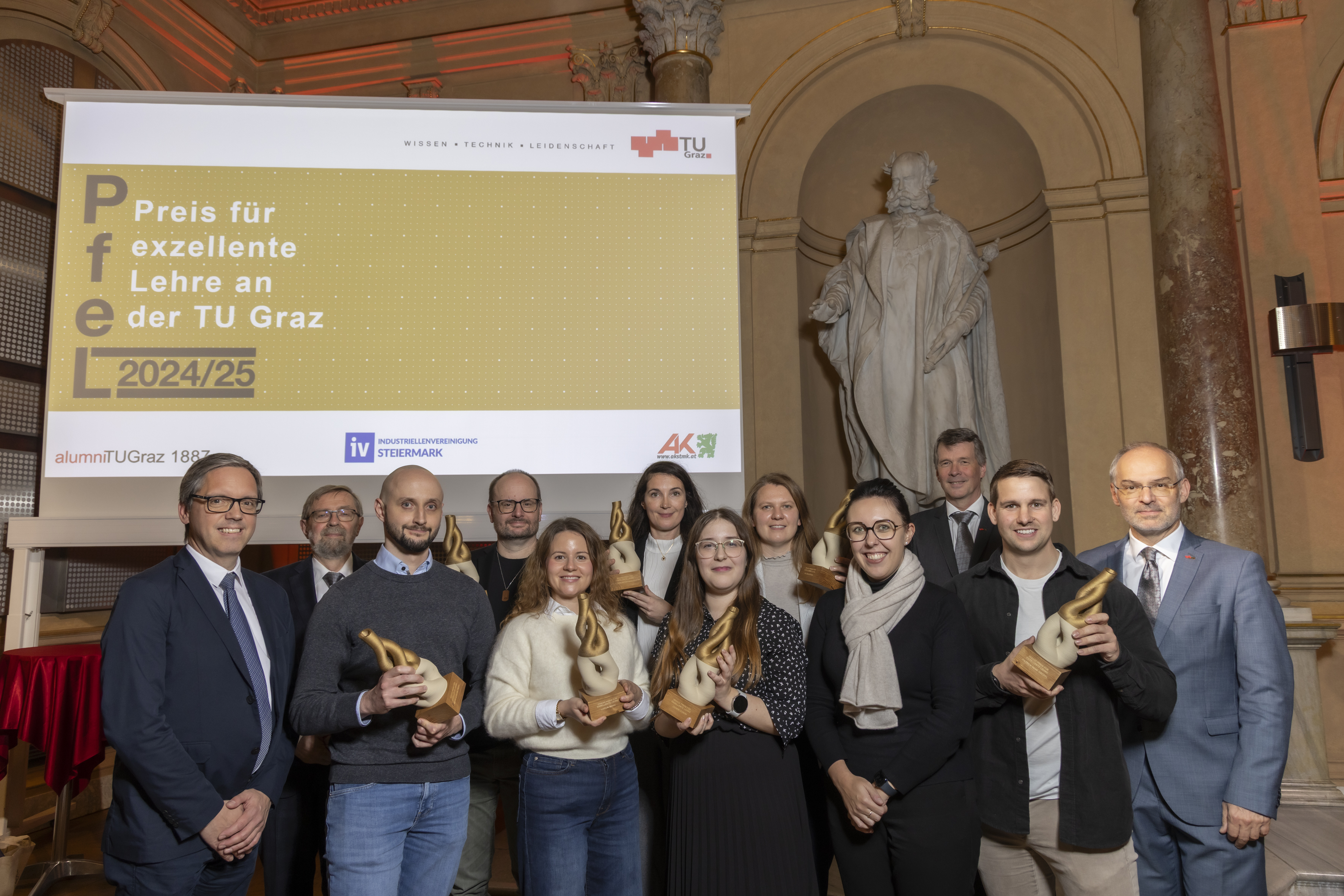 Zwölf Menschen stehen nebeneinnander vor einem Bildschirm und einer Statue.