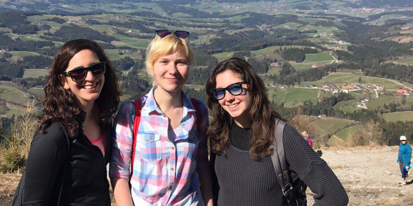 Drei junge Frauen stehen am Gipfel eines Berges und lächeln in die Kamera. Im Hintergrund ist eine weitläufige Landschaft zu sehen.