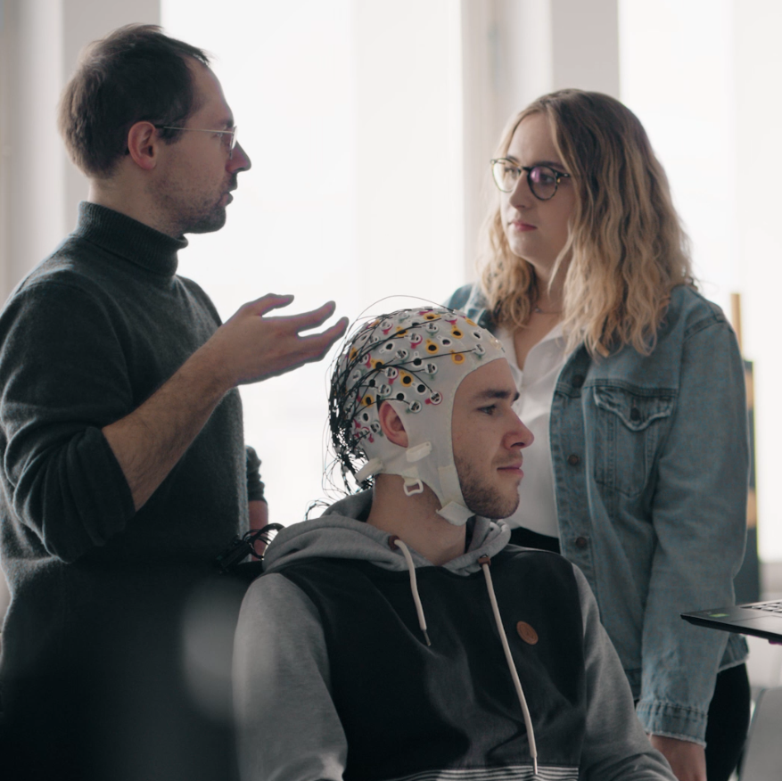3 Personen, eine trägt eine EEG Haube