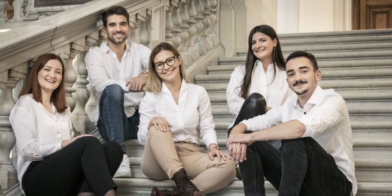 5 Studierende sitzen auf einer Treppe und lächeln in die Kamera